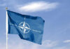 Blue NATO flag with white emblem under cloudy sky.