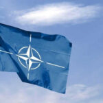 Blue NATO flag with white emblem under cloudy sky.