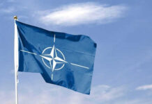 Blue NATO flag with white emblem under cloudy sky.