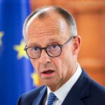 Germany’s Refugee Plan: 80% Must Go! A man in a suit with glasses, looking serious in front of an EU flag