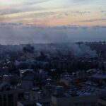 A city skyline at sunset with smoke rising from buildings