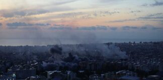 A city skyline at sunset with smoke rising from buildings
