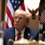 Iran Denies Talks, Trump Claims Progress Man in suit sitting with flag background and eagle decoration