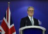 Man in suit at a podium, British flag behind.