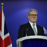 Man in suit at a podium, British flag behind.