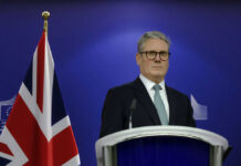 Man in suit at a podium, British flag behind.