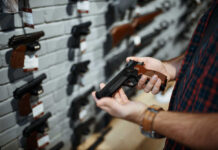 Person holding a handgun in a gun shop with various firearms displayed on the wall