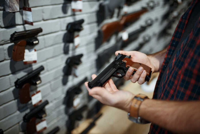 shutterstock_1668328987.jpg Person holding a handgun in a gun shop with various firearms displayed on the wall