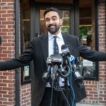 Man in a suit speaking at a press conference with microphones in front of him