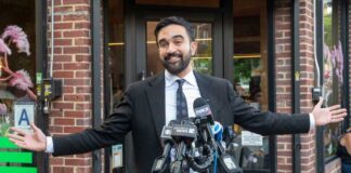 Man in a suit speaking at a press conference with microphones in front of him