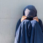 Person in dark blue clothing sitting against a wall with face covered