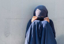 Person in dark blue clothing sitting against a wall with face covered