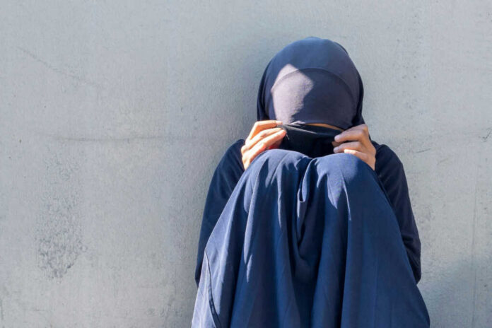 Person in dark blue clothing sitting against a wall with face covered