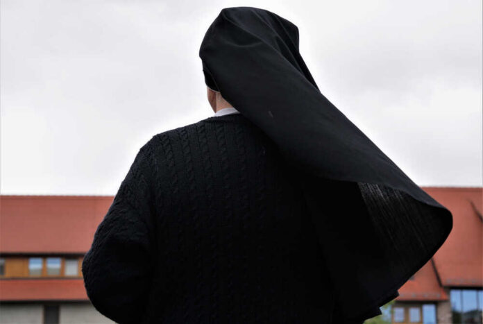 A nun in black attire standing outdoors with a cloudy sky in the background