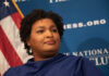 A woman with curly hair speaking at a podium in front of a blue backdrop