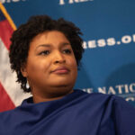 A woman with curly hair speaking at a podium in front of a blue backdrop