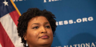 A woman with curly hair speaking at a podium in front of a blue backdrop
