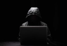 A hooded figure sitting in front of a laptop in a dark setting