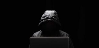 A hooded figure sitting in front of a laptop in a dark setting