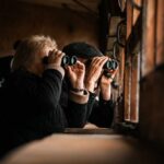 Two individuals using binoculars to observe nature through a window