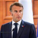 A political leader speaking at a press conference with flags in the background