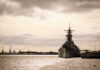 Battleship BOONDOGGLE? Lawmakers Demand Full Design FIRST A naval ship docked in a harbor with industrial cranes in the background under a cloudy sky