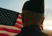 300,000 Veterans Locked Out of Benefits They’re Owed A veteran in uniform standing in front of an American flag at sunset