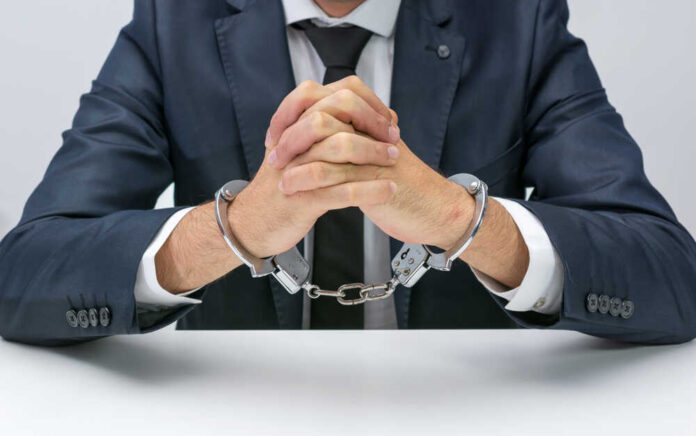 A man in a suit with hands handcuffed together on a table
