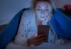 Woman under a blanket looking at a smartphone in bed