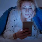 Woman under a blanket looking at a smartphone in bed