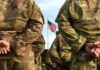 Brittle Arsenal? AI Begins the Audit Soldiers in camouflage stand with hands behind backs facing a U.S. flag