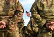 Brittle Arsenal? AI Begins the Audit Soldiers in camouflage stand with hands behind backs facing a U.S. flag