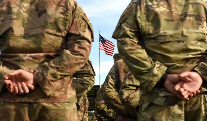 shutterstock_1507031819 Soldiers in camouflage stand with hands behind backs facing a U.S. flag