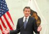 man in suit speaking at podium between U.S. and state flags
