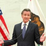 man in suit speaking at podium between U.S. and state flags