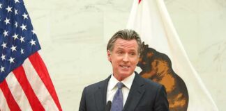 man in suit speaking at podium between U.S. and state flags