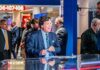 MAGA Infighting Over Tucker Tease news anchor at a studio desk with crew and onlookers