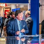 news anchor at a studio desk with crew and onlookers