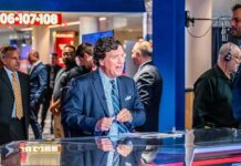MAGA Infighting Over Tucker Tease news anchor at a studio desk with crew and onlookers