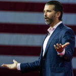 Man speaking in front of American flag backdrop.
