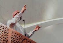 Speaker in traditional attire gesturing from a carved wooden podium