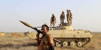 Soldier with RPG launcher walking past armored vehicle with troops