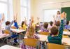 Students raise hands in a classroom while a teacher stands at the front
