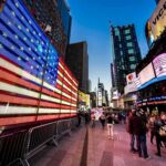 Mayor Hijacks July 4th — Fury Over NYC Speech American flag LED display in a busy city square at dusk