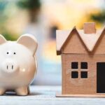 California Housing Math Just Broke Piggy bank beside cardboard house model on a table