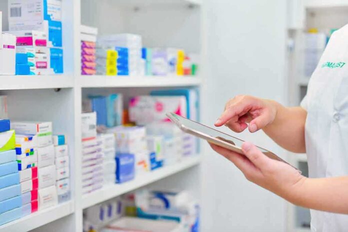 shutterstock_1270720027 Pharmacist using a tablet in front of stocked pharmacy shelves