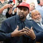 Kanye West speaking while Jim Brown sits nearby in a crowded room